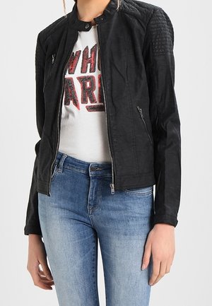 Vrouw die een zwarte jas met ritssluiting draagt met doorgestikte schouders, een wit grafisch T-shirt en een blauwe spijkerbroek, staand tegen een effen achtergrond.