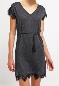 Robe grise à col en V avec des manches courtes, présentant une bordure en dentelle noire au niveau de l'ourlet et des manches, ainsi qu'une taille nouée avec des accents de frange.