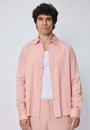 Homme aux cheveux bouclés foncés portant une chemise rose clair ouverte sur un débardeur blanc et un pantalon rose assorti sur un fond uni.