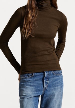 Personne portant un pull à col roulé côtelé marron et un jean en denim bleu, les mains glissées derrière le dos sur un fond blanc uni.
