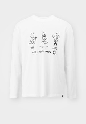 Chemises blanches à manches longues avec des imprimés graphiques, jeans cargo bleu clair avec poches, pantalons gris foncé, lunettes de soleil noires et baskets.