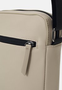Beige syntetisk veske med teksturert overflaten, svart glidelås og flat stropp. Inkluderer en glidelåslomme med metallhardware.