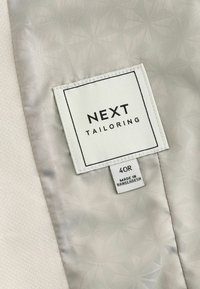 Étiquette en tissu beige clair, arborant la marque "NEXT TAILORING", taille "40R" et "FABRIQUÉ AU BANGLADESH" en impression noire.