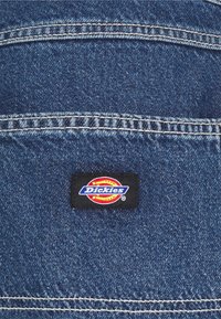 Blue denim fabric with visible stitching details. Black rectangular label with the white and red "Dickies" logo at the top center.