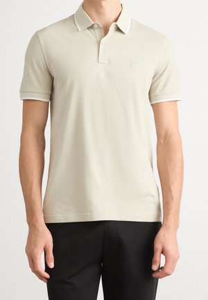 Mann trägt ein beiges Kurzarm-Poloshirt mit weißem Besatz an Ärmeln und Kragen, kombiniert mit schwarzen Hosen, steht vor weißem Hintergrund.