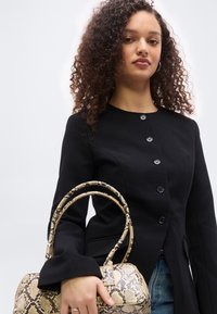 Γυναίκα με σγουρά μαλλιά φοράει μαύρο σακάκι με κουμπιά και κρατά μια τσάντα σε χρώμα ταμπά με σχέδιο φίδι, πάνω σε απλό φόντο.