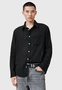 Sort langærmet skjorte i et struktureret stof, med spidse kraver, hvide knapper og en brystlomme, iført med grå jeans og et sort bælte.