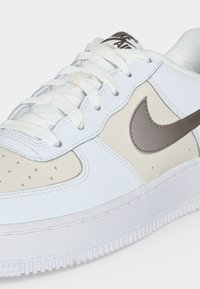 Λευκό και μπεζ Nike Air Force 1 αθλητικό παπούτσι σε κοντινό πλάνο, που δείχνει το γκρι λογότυπο Nike, τα κορδόνια και την υφή της σόλας.