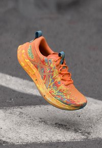 Chaussure de sport orange avec une tige texturée et à motif, ornée d'accents bleus et d'une semelle épaisse et rembourrée, en l'air sur un fond gris.