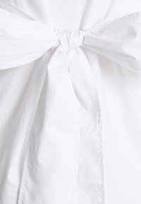 Vitt tyg med en knutdetalj i mitten, med mjuk textur och slät ytfinish; designen betonar enkelhet och elegans.