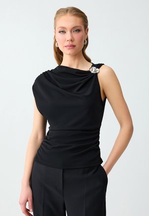 Femme aux cheveux blonds portant un haut noir sans manches avec un col drapé et une broche florale argentée, associée à un pantalon noir, sur un fond uni.