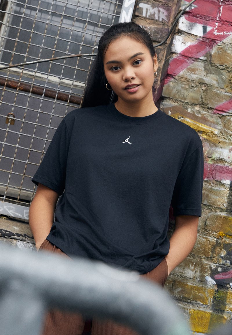 T-shirt en coton noir à manches courtes, avec un logo blanc sur la poitrine. L'arrière-plan comprend un mur en pierre texturée avec des graffitis.
