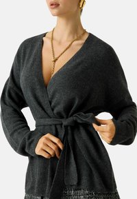 Maglione avvolgente grigio realizzato in un tessuto morbido e strutturato con cintura annodata in vita. Presenta una profonda scollatura a V e maniche lunghe per una vestibilità comoda e rilassata.