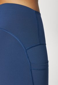 Mørkeblå treningstights laget av elastisk stoff, med flate sømmer og en liten sidelomme for oppbevaring.
