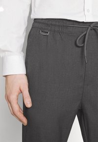 Pantaloni grigi con elastico in vita e cordino, dotati di tasca laterale, caratterizzati da un piccolo etichetta "ONLY & SONS". Il tessuto ha una texture liscia e un motivo minimale.