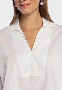 Blouse blanche avec un col cranté et un décolleté en V. Fabriquée en tissu lisse, présentant un design épuré avec des accents minimalistes.