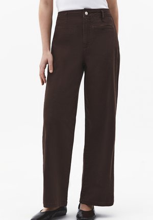 Osoba ubrana w ciemnobrązowe spodnie z wysokim stanem i szerokimi nogawkami, biały top i czarne baleriny, stojąca na tle białego tła.