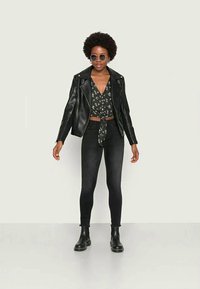 Jaqueta de motoqueiro de couro preto sobre uma blusa floral com laço na frente, combinada com jeans skinny pretos e botas de cano curto pretas. Óculos de sol circulares usados.