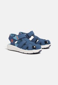 Siniset sandaalit, joissa on suljettu varvas ja viilletty muotoilu. Valmistettu synteettisestä materiaalista, jossa on tarranauhat ja valkoinen pohja, jossa on kuvioitu pinta.