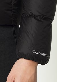Gros plan sur une main portant une manche de doudoune noire avec le logo "Calvin Klein Jeans" près du bord élastique.