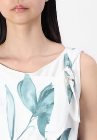 Blouse sans manches en tissu blanc avec un imprimé floral vert. Présente un détail de nœud à l'épaule et une texture douce.