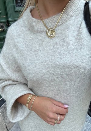 Beige stickad tröja med vida ärmar, prydd med ett guldknut-halsband, flera guldarmband och silvriga ringar på en hand med ljusrosa naglar.