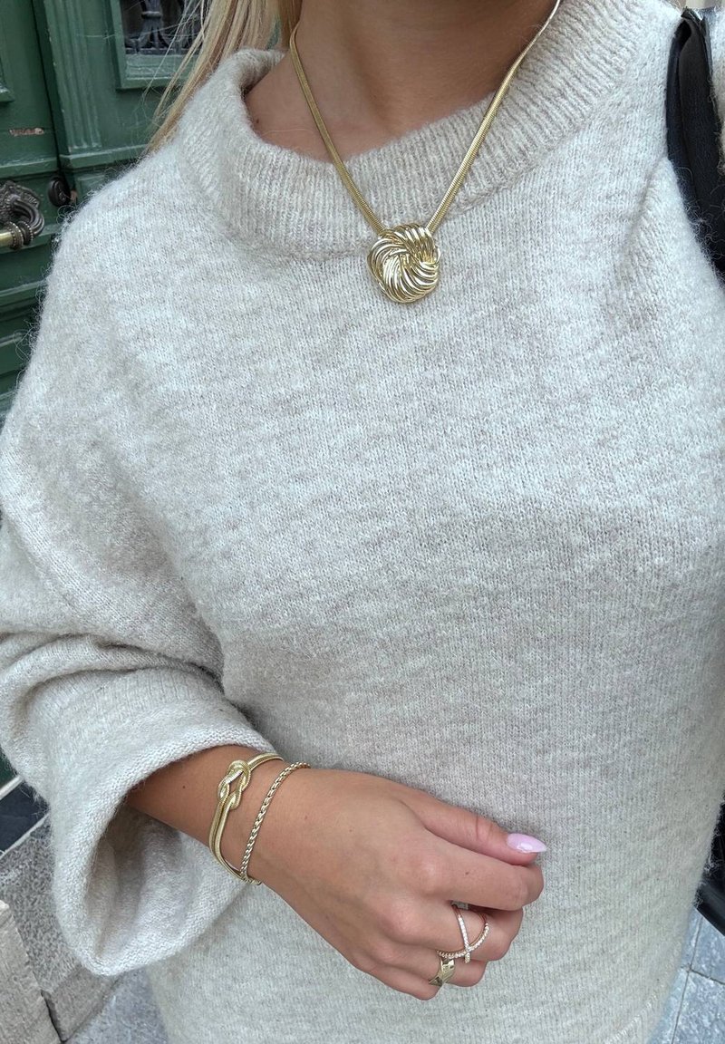 Beige stickad tröja med vida ärmar, prydd med ett guldknut-halsband, flera guldarmband och silvriga ringar på en hand med ljusrosa naglar.