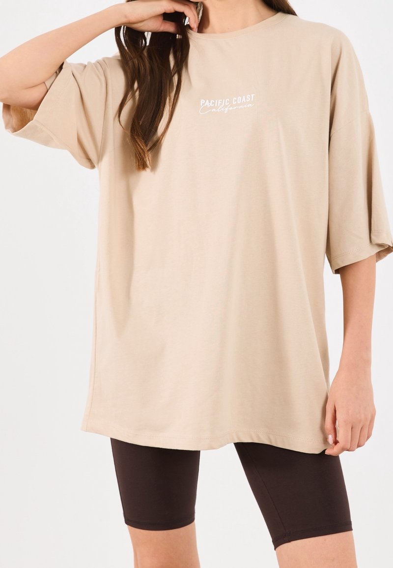 Personne portant un t-shirt beige oversize avec le texte "Pacific Coast California" et un short noir ajusté sur fond blanc.