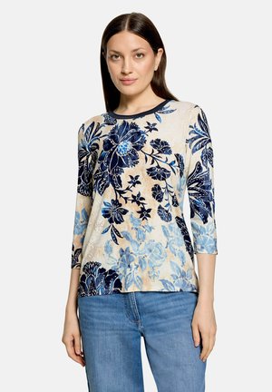 Femme portant un haut à manches longues à motif floral dans des tons de bleu et beige, associé à un jean bleu, debout devant un fond uni.