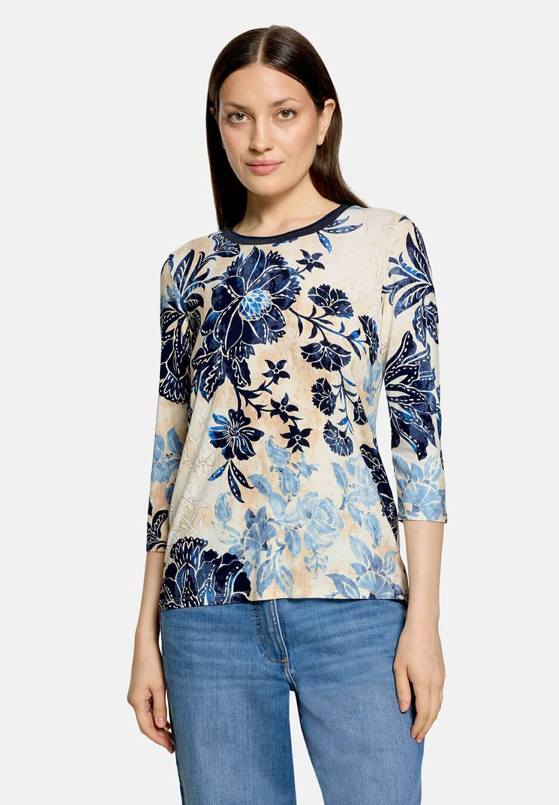 Femme portant un haut à manches longues à motif floral dans des tons de bleu et beige, associé à un jean bleu, debout devant un fond uni.