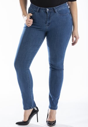 Jean skinny bleu en denim, avec une fermeture à bouton, cinq poches et une coupe classique. Porté avec des chaussures à talons hauts noires.