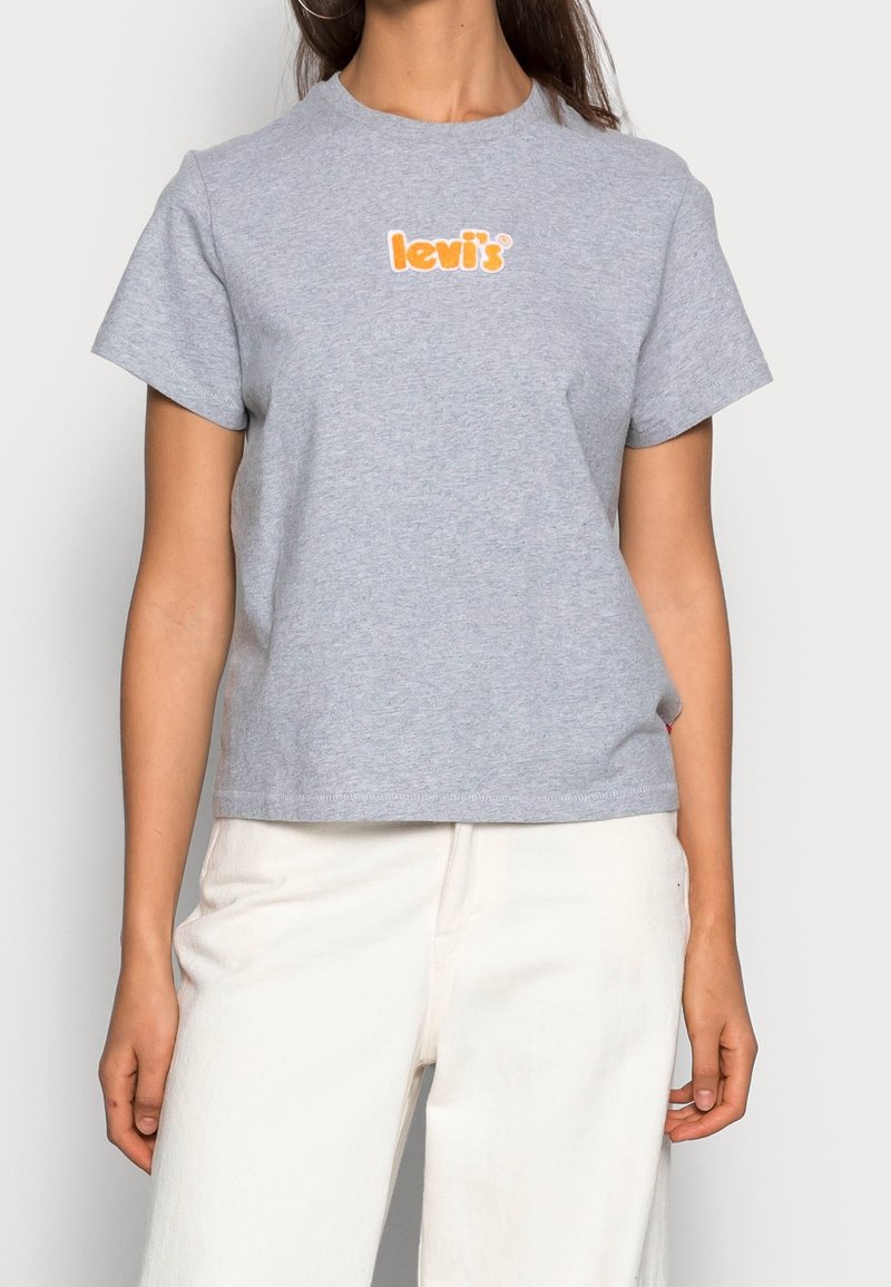 Nainen, joka pitää yllään harmaata lyhythihaista Levi's-t-paitaa, jossa on oranssi logo rinnassa, yhdistettynä korkeavyötäröisiin valkoisiin housuihin yksinkertaista taustaa vasten.