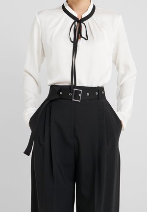 Nainen, joka on pukeutunut valkoiseen paitapuseroon mustilla kaulanauhoilla ja korkeavyötäröisiin mustiin leveälahkeisiin housuihin, joissa on hopealukollinen vyö, kädet taskuissa.