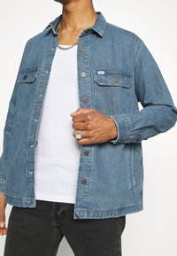 Mies, joka käyttää vaaleansinistä denim Lee-takkia valkoisen ribbipaidan päällä, mustia farkkuja, hopeaketjuneuletta ja hopeasormusta oikeassa kädessä.