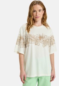 White t-shirt featuring a gold floral design across the chest, short sleeves, and a relaxed fit, paired with light green pants.