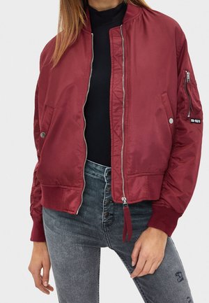 Woman wearing a partially zipped maroon bomber jacket over a black top, paired with high-waisted light gray jeans with button fly details.