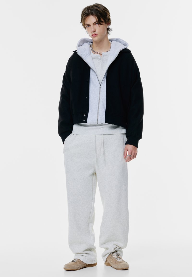 Schwarze Cropped-Jacke über einem grauen Zip-Hoodie, hellgrauem Sweatshirt und passenden Jogginghosen. Helle Sneakers mit Schnürdetails.