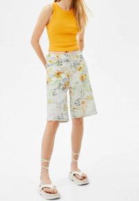 Top sin mangas ajustado amarillo combinado con pantalones cortos de estampado floral multicolor. Los pantalones cortos tienen un corte holgado, cierre de botón y bolsillos. Sandalias de plataforma blancas.