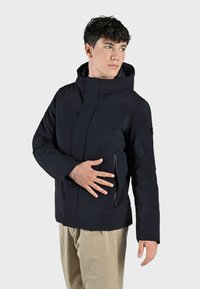 Giacca impermeabile blu navy con colletto alto e cappuccio, dotata di tasca laterale con zip e superficie liscia e opaca.
