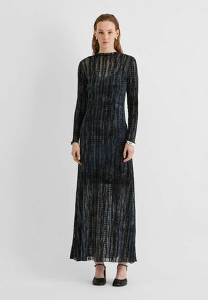Femme portant une longue robe noire transparente à motifs avec des manches longues et des chaussures noires à bride à la cheville, debout devant un fond clair uni.