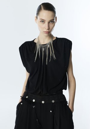 Kobieta z zaczesanymi do tyłu włosami, ubrana w czarny top bez rękawów, szeroki pas z srebrnymi monetami i długi srebrny łańcuch z wiszącymi frędzlami.