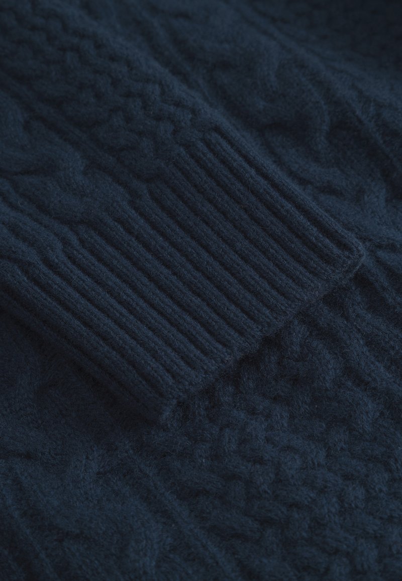 Marineblauer Strickpulloverstoff mit geripptem Bund und strukturierten Mustern, der in einer Nahaufnahme die samtige, wollähnliche Textur zeigt.