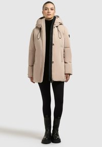 Giacca puffer beige con cappuccio, caratterizzata da cordini neri, ampie tasche e un design leggermente aderente. Indossata sopra un dolcevita nero.