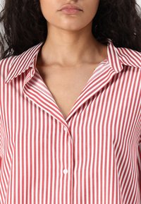 Camisa riscada em vermelho e branco com colarinho clássico, fecho de botão e textura suave de algodão, exibindo riscas verticais e um corte descontraído.