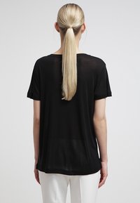 Mujer con una larga coleta rubia que lleva una blusa negra holgada de manga corta y pantalones blancos, vista desde atrás contra un fondo liso.