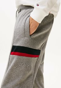 Pantalones de chándal grises con bolsillos laterales, una franja horizontal negra y roja, y un logo en relieve. Hechos de un material suave y texturizado.