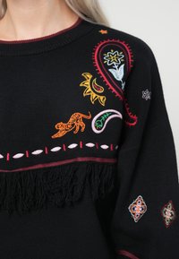 Μαύρο πουλόβερ με πολύχρωμες κεντημένες σχεδιάσεις, συμπεριλαμβανομένων ενός τίγρη, λουλουδιών και μοτίβων paisley. Διαθέτει φ fringe στο κάτω μέρος.