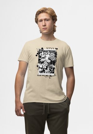 Young man with blonde hair wearing a beige T-shirt featuring a black and white graffiti-style street art design with bold text and abstract shapes.