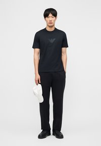 Mann steht, trägt ein schwarzes T-Shirt mit Logo, schwarze Hose, schwarze Schuhe, hält eine weiße Baseballkappe in der linken Hand vor einem einfarbigen Hintergrund.