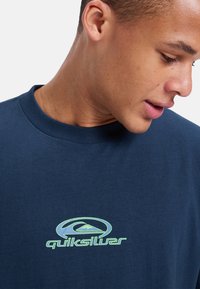 Mørkeblå bomullst-skjorte med rund hals, med en grønn "Quiksilver" logo og fjellgrafikk på brystet.